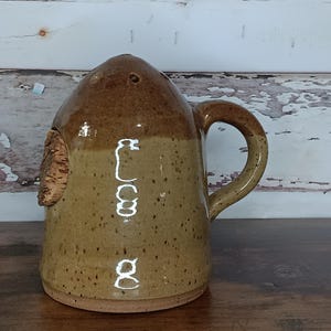 May include: A brown and tan ceramic salt shaker with a cork stopper and a handle. The shaker has a speckled glaze and a light brown glaze on the top half.