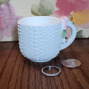 May include: A white, textured teacup with a floral handle. The cup has a bumpy surface and is decorated with pink and green flowers. A silver ring and a coin are next to the cup. The teacup is approximately 3 inches tall.