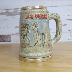 May include: A ceramic beer stein with a beige and brown color scheme. It features a depiction of the Las Vegas skyline with the Golden Nugget Casino and the Horseshoe Casino. The stein has a grapevine-shaped handle.