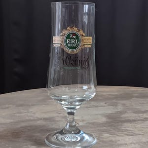 May include: A clear Erlkönig beer glass with a stem and a round base. The glass has a logo with the text "ERL BRAU" and the word "Erlkönig" in script. The glass is on a wooden surface.