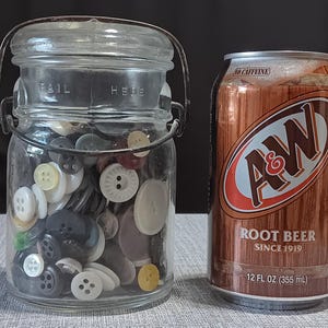 May include: A clear glass jar filled with various buttons in white, black, and yellow, next to a brown A&W Root Beer can. The jar has a metal handle and lid. The can is 12 fl oz (355 mL) and has the text "NO CAFFEINE".