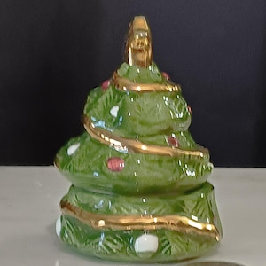 May include: A ceramic Christmas tree figurine. The tree is green with gold trim, red ornaments, and a gold star on top. White accents are visible on the base. The tree is on a white surface.