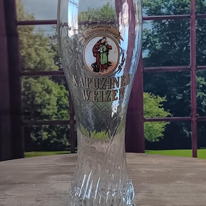 May include: A tall, clear glass with a textured base and a curved body. The glass features the words "KAPUZINER WEIZEN" and a circular emblem with a figure in a brown robe. The glass is designed for beer.