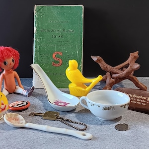 May include: A collection of vintage items including a doll with red hair, a Singer sewing machine instruction manual, a yellow bird figurine, a ceramic spoon, a teacup, and a wooden stand. The instruction manual reads "SINGER SEWING MACHINE 15-86".