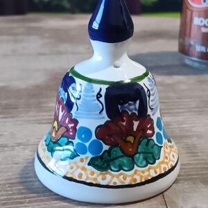 May include: A hand-painted ceramic bell with a dark blue top and a white base. The bell features floral designs in red, blue, and green, with a yellow and black border. The bell is a decorative item.