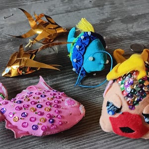 May include: An assortment of handmade fish ornaments in various styles and colors. Two are gold, origami-style fish. One is pink with sequins, another is blue with a yellow fin, and the last is a peach-colored face with a red mouth.