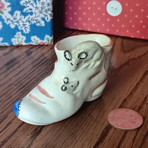 May include: A small, off-white ceramic boot figurine with two mice figures. The boot has red and blue accents. A copper coin is next to the boot. Decorative boxes are in the background.