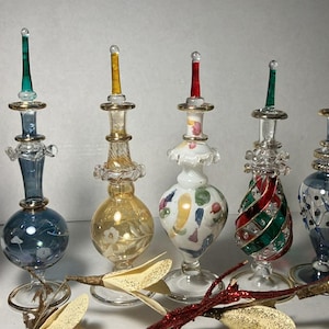 May include: A selection of six glass Christmas tree toppers. Each topper has a different design and colour scheme, with gold accents. The finials are in various colours, including red, green, and blue. The bases are decorative.