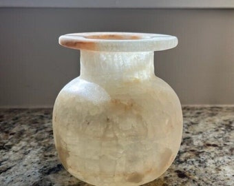 May include: A small, white alabaster vase with a round body and a wide, flat rim. The vase has a slightly translucent quality and shows signs of natural veining.