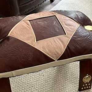May include: A rectangular ottoman featuring a patchwork design of dark brown and beige leather panels. The leather surfaces are embossed with crest-like patterns. The piece is accented with cream-colored piping and circular gold-toned metal hardware.