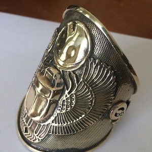 May include: A gold-toned metal bracelet with an intricate design featuring a scarab beetle, wings, and a yin-yang symbol.
