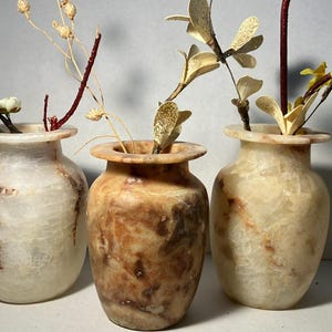 Puede incluir: Tres jarrones decorativos de piedra con arreglos florales. Los jarrones son en tonos crema y marrón, con patrones de vetas únicos. Cada jarrón contiene diferentes flores y ramas secas, agregando un toque natural a la decoración.