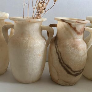 Puede incluir: Cuatro jarrones decorativos de alabastro con tapas y asas. Los jarrones son en tonos crema y beige, algunos con vetas marrones. Un jarrón contiene tallos secos.