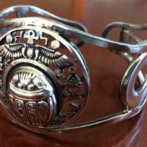 Könnte beinhalten: Silberfarbenes Armreif-Armband mit ägyptischem Design. Das Armband hat ein ovales Medaillon mit einem Skarabäus, Hieroglyphen und geflügelten Elementen. Das offene Design umfasst gebogene Stäbe und dekorative Ausschnitte.