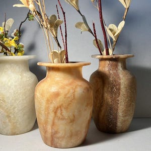 Puede incluir: Tres jarrones decorativos de piedra en tonos crema, beige y marrón. Cada jarrón contiene arreglos florales secos. Los jarrones tienen una forma de urna clásica y se exhiben sobre un fondo neutro.