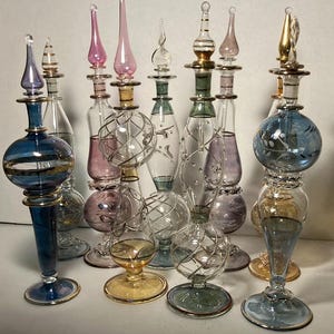 May include: A collection of decorative glass perfume bottles in various shapes and colours. The bottles feature intricate designs, including spherical and teardrop shapes, with gold accents and colourful stoppers. The colour palette includes blue, pink, and clear glass.
