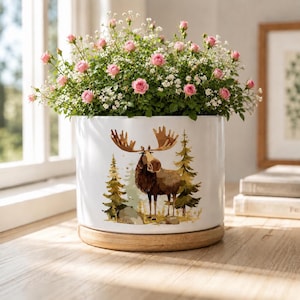 May include: A white cylindrical ceramic planter with a wooden base, featuring a graphic illustration of a brown moose standing among green pine trees. The planter is filled with a lush arrangement of small pink roses and white baby's breath flowers.
