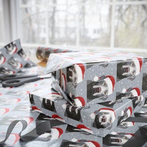 May include: Wrapped gifts with festive wrapping paper featuring a repeating pattern of dogs wearing Santa hats. The gifts are stacked on a table with scissors, ribbon, and additional wrapping paper visible in the background. The paper is gray with white snowflakes.