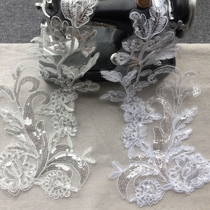 Może przedstawiać: Dwa białe aplikacje z koronki kwiatowej z srebrnymi cekinami. Aplikacje znajdują się na jasnoszarym tle z tkaniny.