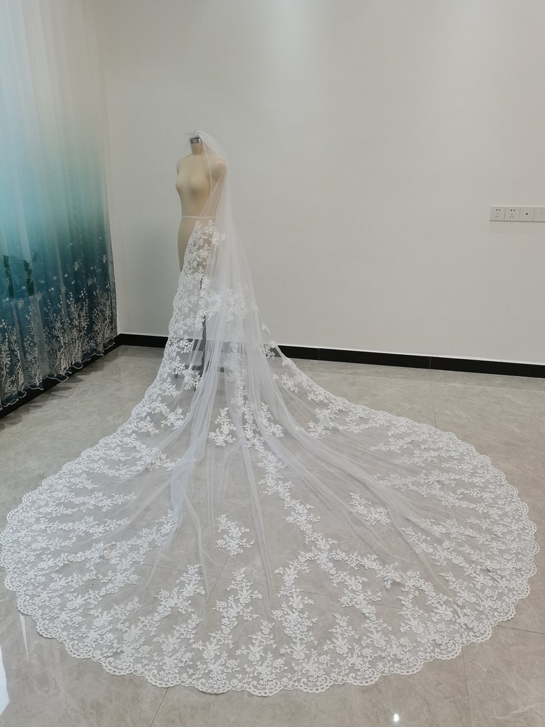 Puede incluir: Un velo de novia blanco con delicado bordado de encaje floral. El velo es largo y fluido, con un borde festoneado.