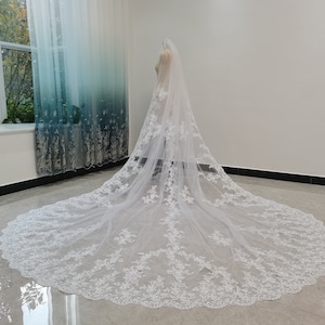 Puede incluir: Un velo de novia blanco con delicados detalles de encaje y un borde festoneado. El velo es largo y fluido, con una longitud de catedral.