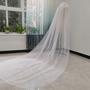 May include: A long, white tulle bridal veil with a delicate, flowing design. The veil is draped over a mannequin and is shown against a white background.