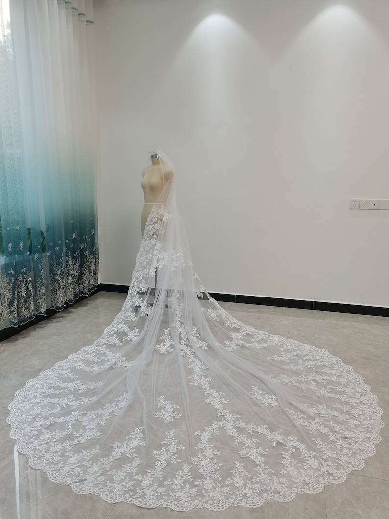 Puede incluir: Un velo de novia blanco con delicado bordado de encaje y un borde festoneado. El velo se muestra en un maniqu&iacute; en una habitaci&oacute;n blanca.