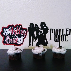 May include: Three cupcakes decorated with black frosting and topped with black and white silhouette cake toppers featuring the band Motley Crue.