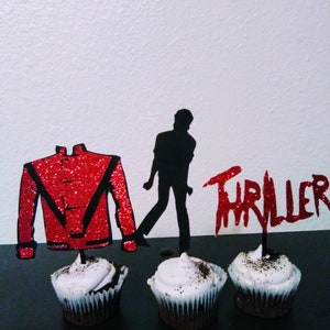 May include: Three cupcakes decorated with black sprinkles and topped with red and black paper cutouts. The cutouts are a red jacket, a silhouette of a person, and the word "Thriller".