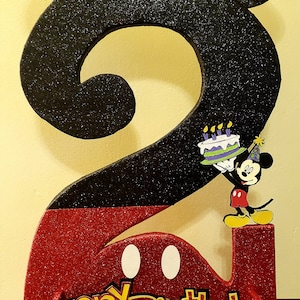 May include: A red and black glitter number two with Mickey Mouse ears and a Mickey Mouse figure holding a birthday cake. The number two is decorated with white polka dots. The words "Happy Birthday" are written in yellow letters with black outlines.