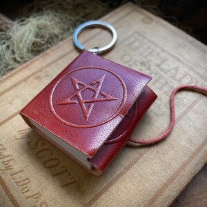 May include: A small, brown leather-bound journal with a pentagram embossed on the front cover. The journal is attached to a silver key ring and has a red cord.