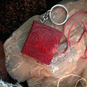 May include: A red leather journal with a tree of life design and a silver key chain. The journal is closed and has a red cord attached to it.