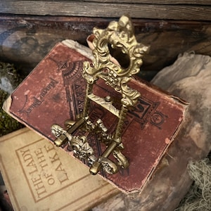 K&ouml;nnte beinhalten: Ein dekorativer St&auml;nder aus goldenem Metall mit runder Oberseite und drei Beinen. Der St&auml;nder ist mit filigranen floralen Details verziert und hat eine Vintage-&Auml;sthetik.