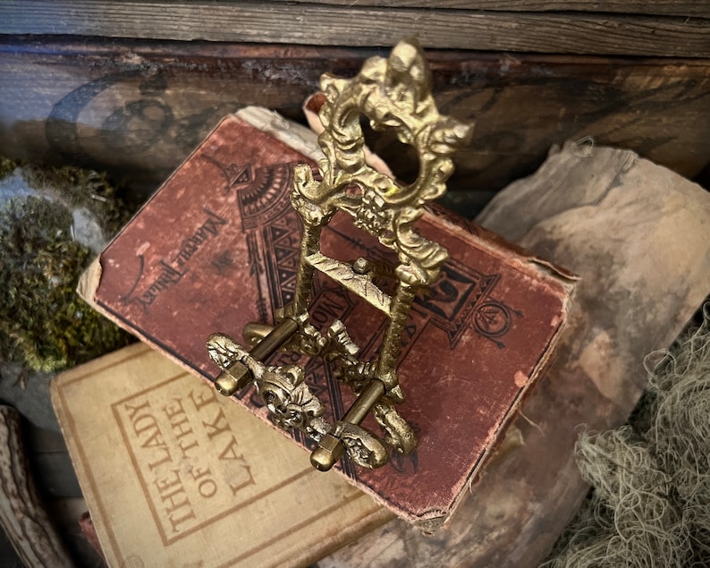 K&ouml;nnte beinhalten: Ein dekorativer St&auml;nder aus goldenem Metall mit runder Oberseite und drei Beinen. Der St&auml;nder ist mit filigranen floralen Details verziert und hat eine Vintage-&Auml;sthetik.