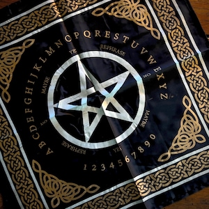 May include: Black divination cloth with a white pentagram in the center, surrounded by the alphabet, numbers, and phrases like "yes," "no," and "maybe." Gold Celtic knotwork borders the edges, creating a mystical aesthetic.