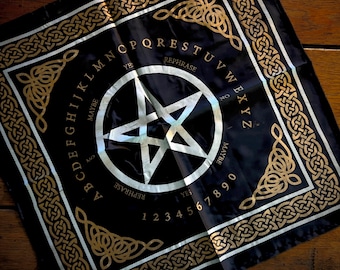 May include: Black divination cloth with a white pentagram in the center, surrounded by the alphabet, numbers, and phrases like "yes," "no," and "maybe." Gold Celtic knotwork borders the edges, creating a mystical aesthetic.