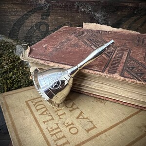 May include: A silver bell with a pentagram design on an antique book. The book has a brown leather cover with embossed lettering and is titled "The Law of the Lady".