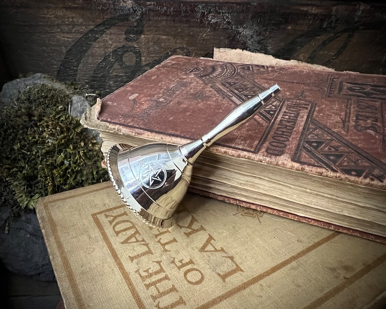 May include: A silver bell with a pentagram design on an antique book. The book has a brown leather cover with embossed lettering and is titled "The Law of the Lady".