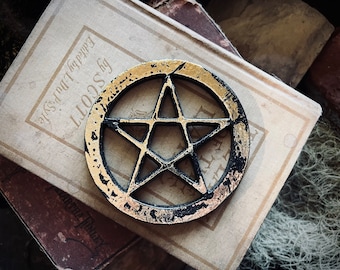 May include: A black metal pentagram with gold accents, resting on top of a stack of old books. The pentagram is a five-pointed star, a symbol often associated with witchcraft and paganism.