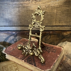K&ouml;nnte beinhalten: Ein kleiner, verzierter, goldfarbener Metall-Staffelei-St&auml;nder mit einem dekorativen floralen Design. Der St&auml;nder steht auf einem Stapel alter B&uuml;cher.