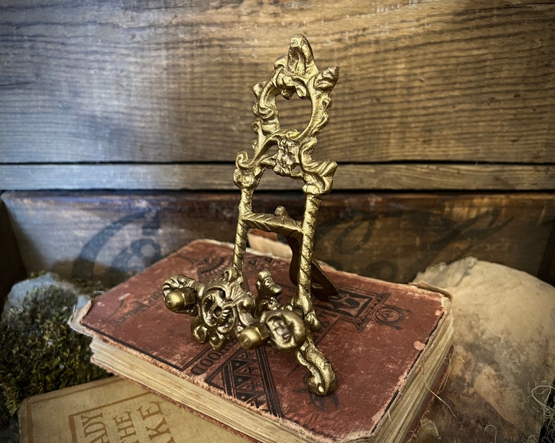 K&ouml;nnte beinhalten: Ein kleiner, verzierter, goldfarbener Metall-Staffelei-St&auml;nder mit einem dekorativen floralen Design. Der St&auml;nder steht auf einem Stapel alter B&uuml;cher.