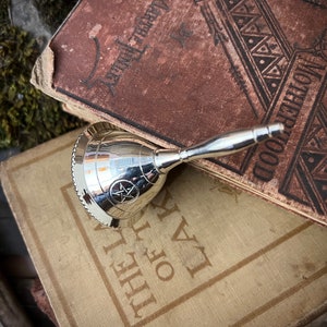 May include: A silver bell with a pentagram design on the front, resting on top of two antique books. The top book has the word "Motherhood" printed on the cover.