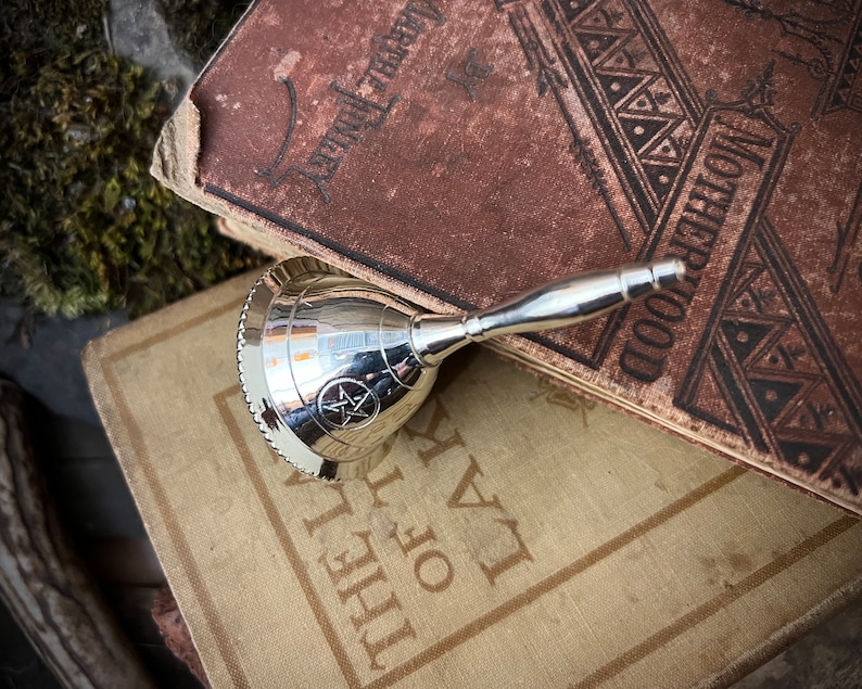 May include: A silver bell with a pentagram design on the front, resting on top of two antique books. The top book has the word "Motherhood" printed on the cover.