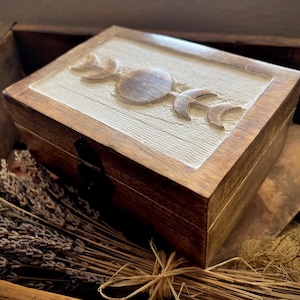 Peut inclure: Une boîte en bois avec un motif de phases lunaires sculpté sur le couvercle. La boîte est marron avec un panneau texturé de couleur claire sur le couvercle. Les phases lunaires sont en relief et ont une finition en bois naturel. La boîte est rectangulaire.