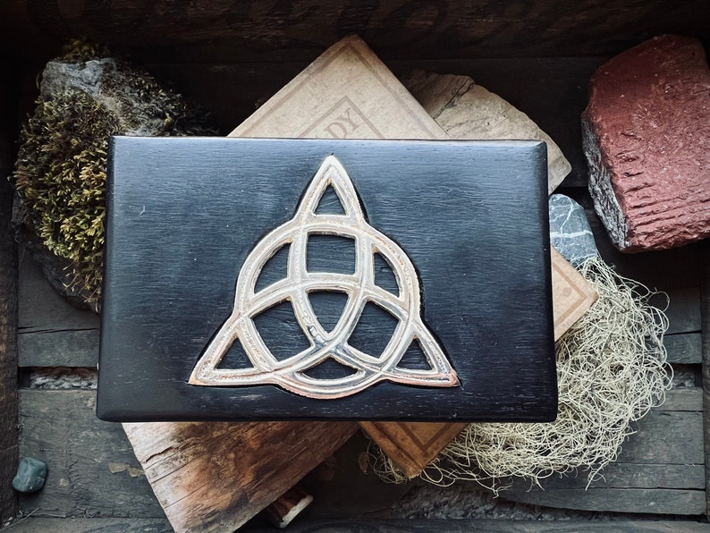 May include: A black wooden box with a silver Celtic knot design on the lid. The box is sitting on top of a stack of old books.