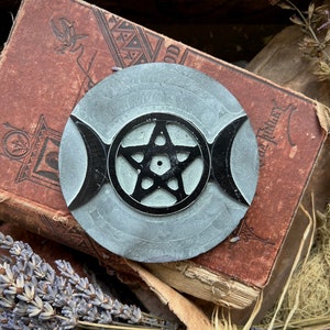 May include: A gray stone incense burner with a pentagram and triple moon design. The burner is on top of a vintage book.