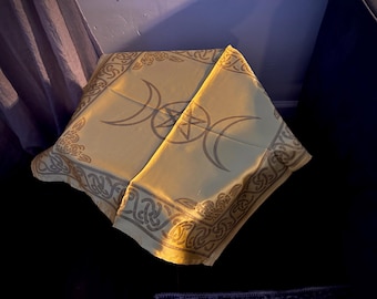 May include: A square, pale yellow fabric cloth featuring a central triple moon and pentagram symbol. The border is decorated with a repeating dark brown Celtic knotwork pattern, with additional knotwork designs in each of the four corners.