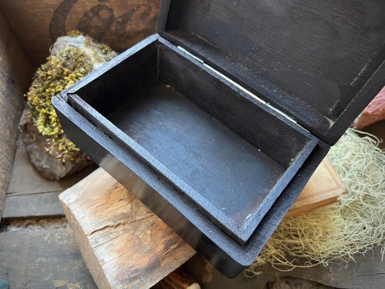 May include: A black wooden box with a hinged lid, open to reveal an empty interior. The box is made of dark wood and has a smooth finish.