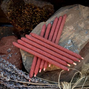 May include: A collection of red incense sticks arranged in a starburst pattern. The incense sticks are a deep red color with a textured surface. The background includes rocks, dried lavender, and other natural elements.