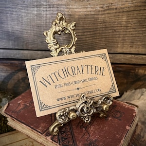 May include: A gold metal decorative stand with a scroll design holds a business card that reads "Witchcrafterie Ritual Tools + Curios + Spell Supplies www.witchcrafterie.com".